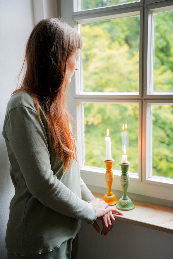 Eine Frau steht vor dem Fenster. neben ihr zwei Kerzen. Sie schaut aus dem Fenster und trägt einen selbstgenähten Schlafanzug.