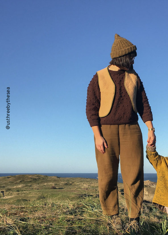 Eine Frau steht in Spanien am Meer und träft die genähte Weste Malva. An der Hand hält sie ein Kind.