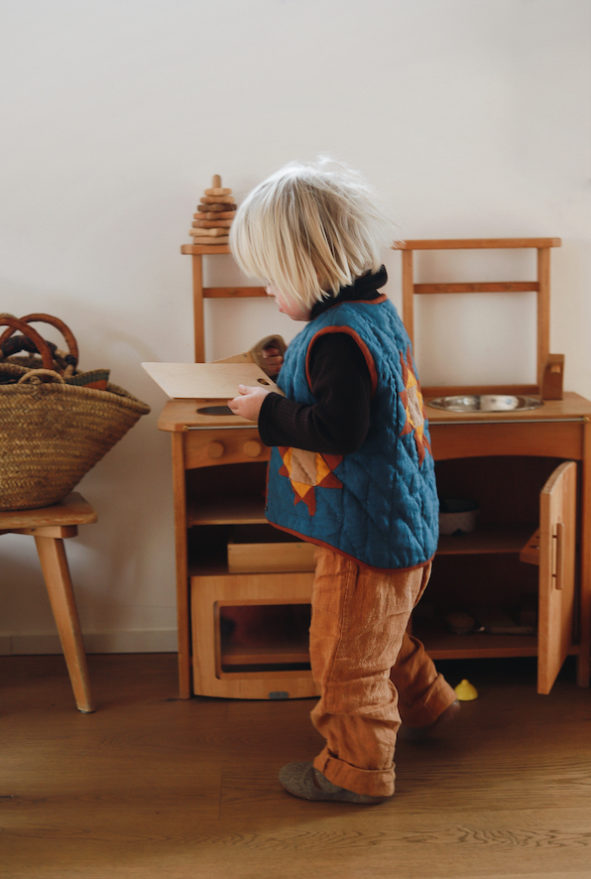 Ein Junge mit Down Syndrom spielt mit einer Holzküche im Wohnzimmer. Er trägt eine gequiltete Weste von Nanna Pattern Design.
